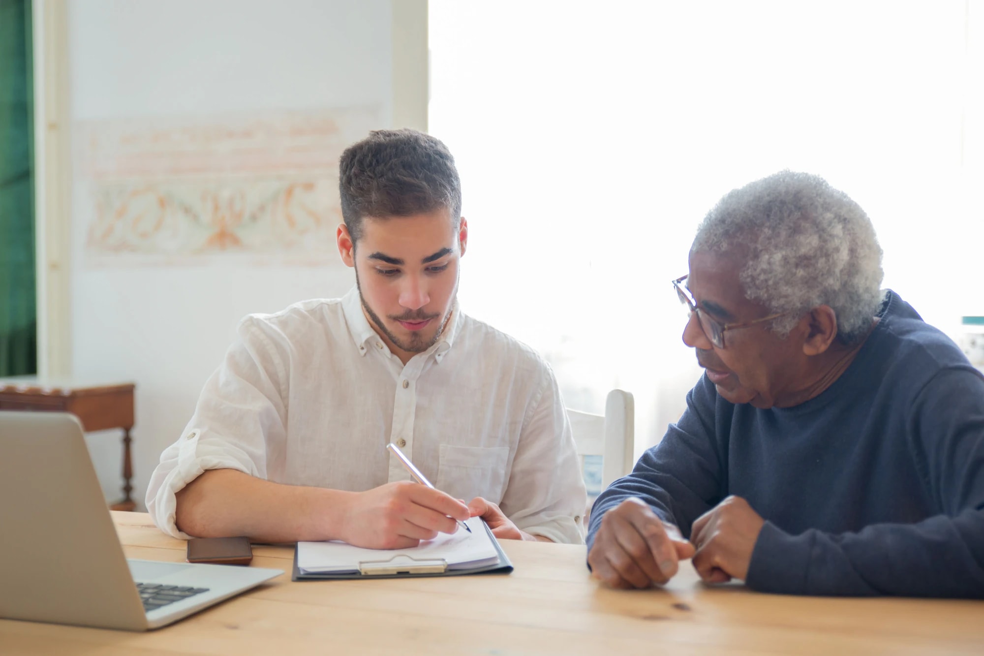 A young man helping an older man fill in paperwork, with a laptop on the table