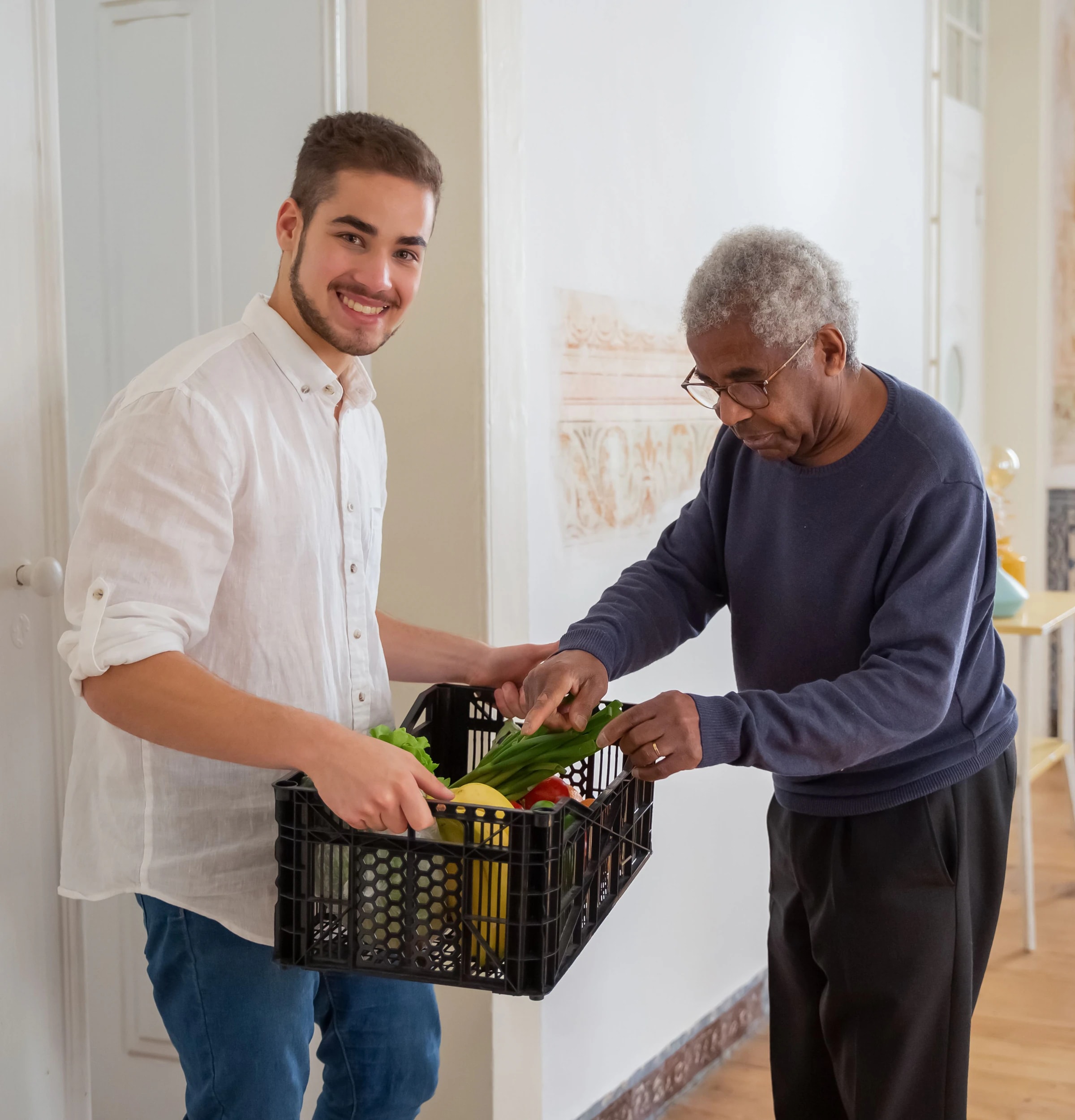 A young man handing a crate of fresh vegetables to an older man at his home