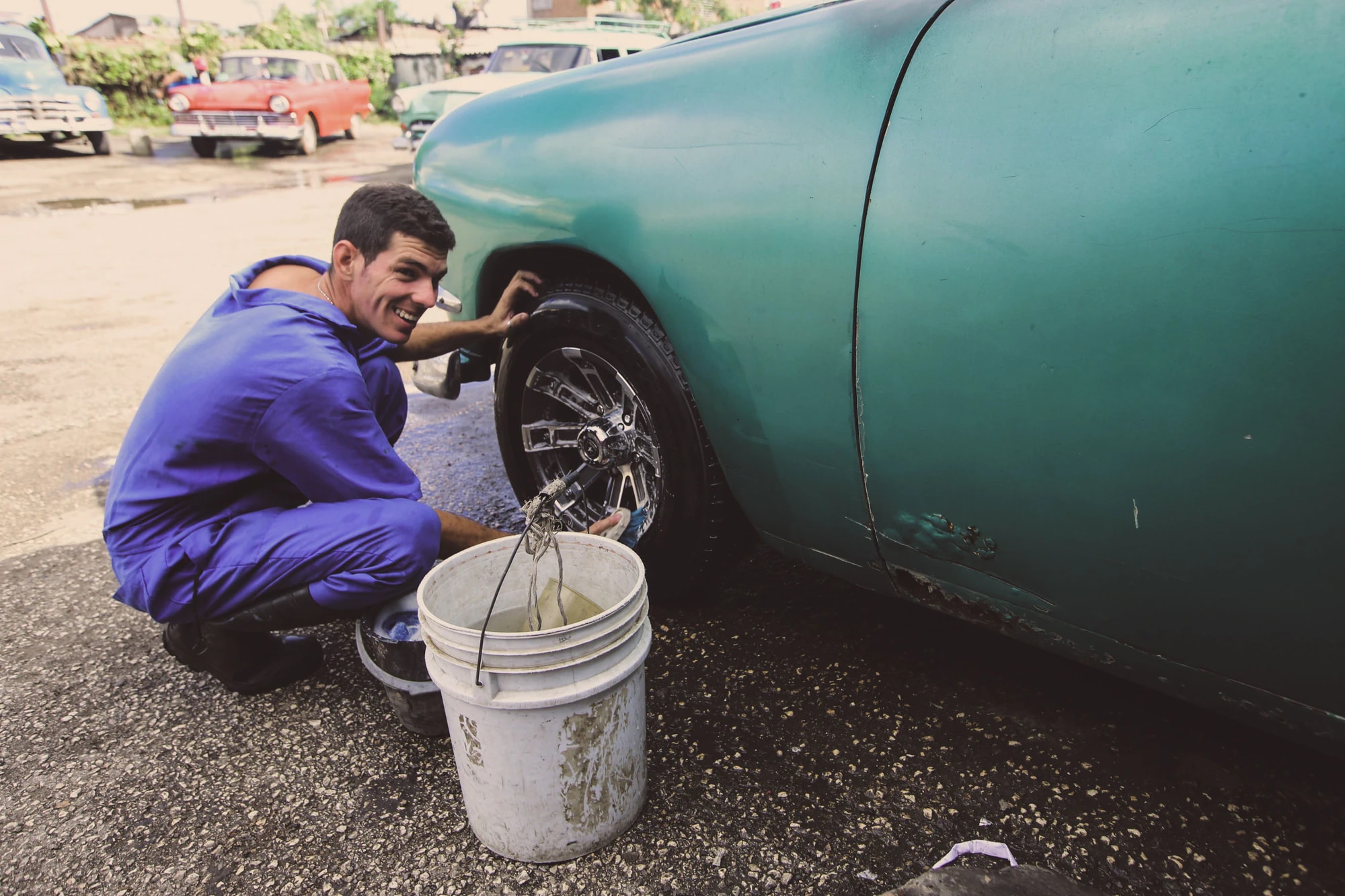 A man smiling as he cleans a car’s wheel with a bucket beside him